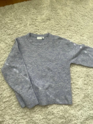 Ljuslila stickad tröja från Vila - Mysig ljuslila stickad tröja från Vila med rund halsringning och ribbade muddar vid ärmslut och nederkant. Tröjan har en enkel, avslappnad passform och är perfekt för lager på lager under kyliga dagar.