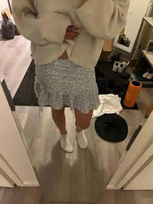 Ljusblå blommig volangkjol - Supersöt ljusblå kortkjol med vitt blommönster och volangkant. Kjolen har snörning i sidan för extra detalj och är perfekt för vår och sommar. Bekväm passform och lätt material som känns luftigt. Perfekt till sommaren