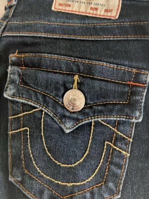 True Religion mörkblå bootcut jeans - Säljer ett par mörkblå jeans från True Religion med ikoniska kontrastsömmar och snygga fickdetaljer med silverknappar. Jeansen har bootcut-modell och klassisk femficksdesign. Perfekta för dig som vill ha en statement look.