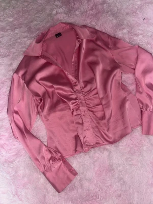 Rosa satinblus med rynkade detaljer - Snygg rosa blus i glansigt satinmaterial med rynkade detaljer framtill. Blusen har långa ärmar med breda manschetter och klassisk krage. Figurnära passform och knäppning framtill ger en trendig look. I storlek XL