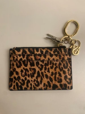 Leopardmönstrad korthållare med nyckelring - Snygg korthållare i leopardmönstrat tyg med dragkedja upptill och fack på framsidan. Utrustad med guldfärgad nyckelring och karbinhake, samt en dekorativ metallcharm. Perfekt för kort och nycklar i ett.