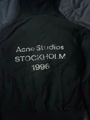 Svart hoodie Acne Studios Stockholm - Svart hoodie från Acne Studios med stor huva och tryck på ryggen där det står 'Acne Studios STOCKHOLM 1996' i vitt. Klassisk passform och ribbade muddar. Perfekt för dig som gillar streetwear och vill ha något stilrent med en touch av svensk design. Kvitto finns