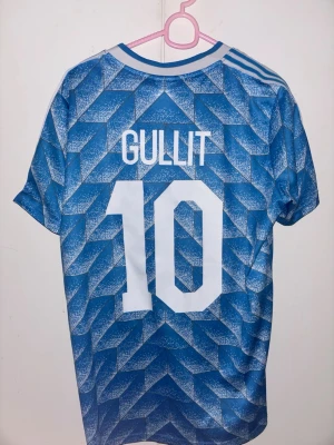 Retro Nederländerna fotbollströja Gullit 10 - Retro blå fotbollströja från Adidas med vit geometriskt mönster, vita ränder på axlarna och trycket 'GULLIT' samt nummer 10 på ryggen. Klassisk V-ringning och broderad logga samt Nederländernas lejonmärke på bröstet. Tröjan är från EM-guldåret 1988. Enligt mig en av de snyggaste retrotröjorna därute. 