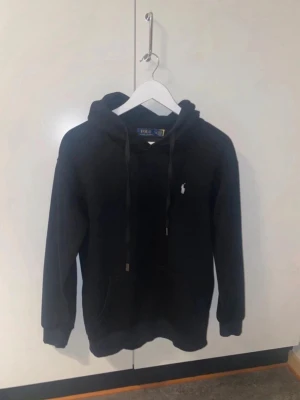 Svart hoodie från Polo Ralph Lauren - Snygg svart hoodie från Polo Ralph Lauren med klassisk vit broderad logga på bröstet. Hoodien har huva med dragsko, magficka och ribbade muddar vid ärmar och nederkant. Tillverkad i mjukt bomullsmaterial för skön känsla.