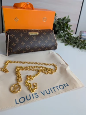 Iouis Vuitton Pochette Eva Monogram 🤎 - Ikonisk väska med guldkedja – Nyskick! ✨ Säljer en fantastiskt elegant väska i klassiskt mörkt mönster. Den har en feminin design med en tillhörande, kraftig guldkedja som pryds av vackra berlocker. Perfekt som både axelremsväska och lyxig clutch för kvällen. Skick: Helt ny och oanvänd. Skyddsplasten sitter kvar på metalldetaljerna framtill. Medföljer: Ljus original-dustbag och den karaktäristiska orangea boxen.