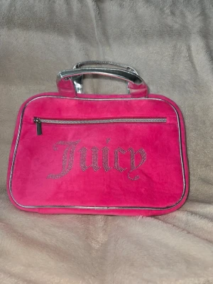 Juicy Couture rosa sminkväska  - Säljer en ikonisk rosa handväska från Juicy Couture i mjuk velour med silverfärgade detaljer och handtag. Väskan har en dragkedja framtill och texten 'Juicy' i glittriga stenar på framsidan. Perfekt för dig som gillar en färgstark och trendig look.