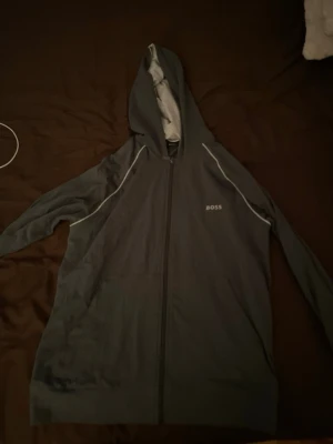 Mörkgrå hoodie från BOSS - Snygg mörkgrå hoodie från BOSS med dragkedja framtill och vit piping längs axlarna. Hoodien har en stor huva med ljusgrå insida och diskret BOSS-logga på bröstet. Perfekt för en chill och stilren look.