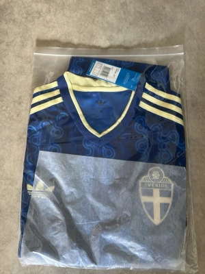 Blå Sverige fotbollströja Adidas - Säljer en blå fotbollströja från Adidas med Sveriges landslagsemblem på bröstet. Tröjan har gula detaljer, V-ringning och klassiska Adidas-ränder på axlarna. Materialet är lätt och andas, perfekt för träning eller match.