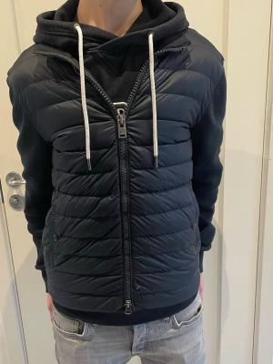 Woolrich cardigan  - Svart cardigan med quiltad framsida och mjuka ärmar samt rygg i sweatshirtmaterial. Jackan har dragkedja framtill, två sidofickor och en huva med vita snören. Perfekt för kalla dagar och riktigt snygg streetstil.
