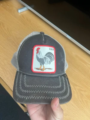 Svart truckerkeps från Goorin Bros - Säljer en svart och vit truckerkeps från Goorin Bros med mesh på sidorna och bak. Framtill finns ett broderat tuppmärke med röd kant och texten 'COCK'. Kepsen har justerbar snapback och coola zickzack-sömmar på skärmen.