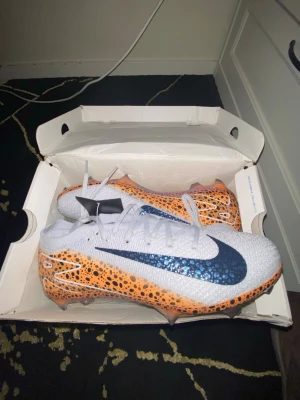 Nike Mercurial fotbollsskor orange/vit - Säljer ett par Nike Mercurial fotbollsskor med vit ovandel och orange sula med svarta leopardfläckar. Skorna har högt skaft och en stor svart Nike-swoosh på sidan. Tillverkade i syntetmaterial för bästa grepp och känsla på planen.