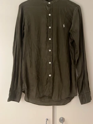 Olivgrön skjorta från Polo Ralph Lauren - Stilren olivgrön skjorta från Polo Ralph Lauren med klassisk rak passform och vita knappar framtill. Skjortan har lång ärm, rundad krage och den ikoniska loggan broderad på bröstet. Perfekt för en clean och avslappnad look.