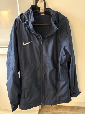 Mörkblå Nike vindjacka med huva - Snygg mörkblå vindjacka från Nike med dragkedja framtill och justerbar huva. Jackan har en minimalistisk design med Nike-logga på bröstet och är tillverkad i lätt, vindtätt material. Perfekt för blåsiga dagar och enkel att matcha med olika outfits.