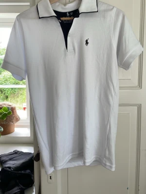 Vit piké från Ralph Lauren - Klassisk vit pikétröja från Ralph Lauren med svart broderad logga på bröstet och svart kant på kragen. Skjortan har korta ärmar och är gjord i mjuk bomull. Perfekt till en clean och stilren look.