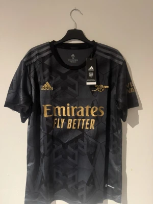 Arsenal bortatröja Adidas svart/guld - Snygg Arsenal bortatröja från Adidas i svart med guldiga detaljer och loggor. Tröjan har geometriskt mönster, rund hals och korta ärmar. Tillverkad i lätt och ventilerande material, perfekt för fotboll eller träning.