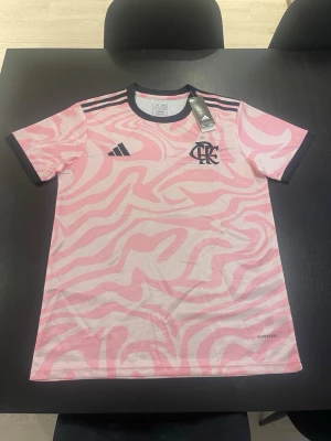 Rosa Flamengo träningströja Adidas - Säljer en rosa och vit träningströja från Adidas med Flamengo-logga och svart stjärna på bröstet. Tröjan har svarta detaljer, rund hals och klassiska Adidas-ränder på axlarna. Tillverkad i lätt och ventilerande material, perfekt för träning eller match.