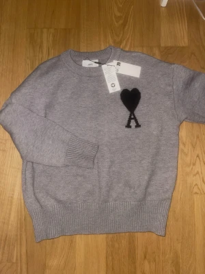 Grå stickad tröja från Ami Paris - Snygg grå stickad tröja från Ami Paris med svart hjärta och bokstaven A broderat på bröstet. Tröjan har rund halsringning, ribbade muddar och är tillverkad i mjukt material. Perfekt för en stilren och trendig look.