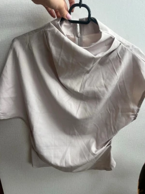 Beige omlottopp med hög krage - Snygg beige topp med omlottkrage och korta ärmar. Toppen har en lös passform och dragkedja i nacken för enkel påklädning. Perfekt för dig som gillar stilrena och moderna plagg.  OANVÄND