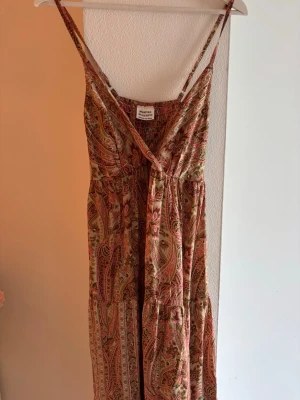 Mönstrad långklänning  - Bohemisk långklänning från Bluevision med smala axelband och omlott-v-ringning. Klänningen har ett paisleyinspirerat mönster i beige, rosa och bruna toner. Perfekt för sommarens alla tillfällen och tillverkad i ett mjukt, luftigt material.