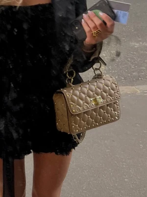 Beige väska med nitar - Säljer en beige quiltad handväska med guldfärgade nitar och kedjerem. Väskan har ett elegant rutmönster, vridlås framtill och detaljer i metall. Perfekt för dig som vill ha en trendig och lyxig accessoar till din outfit.