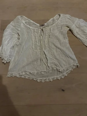 Vit boho-blus med spets från Zara - Romantisk vit blus från Zara med boho-vibbar. Blusen har långa ärmar, dekorativa spetsdetaljer längs kanterna och framtill samt knytning vid halsen. Tillverkad i ett lätt, mjukt tyg som ger en avslappnad look. Ett litet hål vid spetsen i ena ärmen samt en super liten fläck där fram , står ingen storlek men skulle gissa på xs-s 