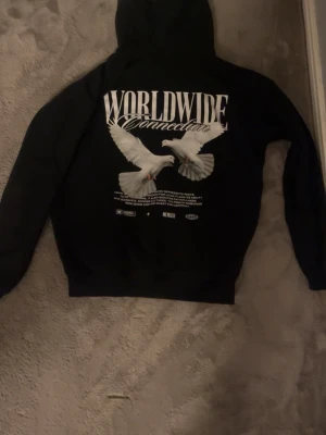 Svart hoodie Worldwide Connection - Svart hoodie med stor vit text 'WORLDWIDE Connection' och två vita duvor tryckta på ryggen. Mindre tryck på bröstet och klassisk känguruficka framtill. Hoodien har huva och ribbade muddar. Snygg streetwear vibe och skön passform.