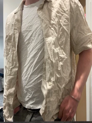 Beige linneskjorta med kort ärm - Säljer en beige skjorta i linne med korta ärmar och klassisk krage. Skjortan har knappar framtill och en bröstficka. Perfekt för varma dagar och enkel att styla över en t-shirt.