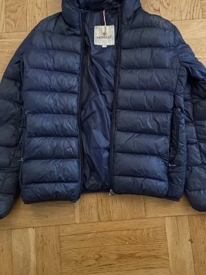 Mörkblå dunjacka från Moncler - Snygg mörkblå dunjacka från Moncler med huva och quiltad design. Jackan har två sidofickor med dragkedja och en hög krage. Klassisk Moncler-logga på ärmen och lätt glansigt material som ger en exklusiv känsla. Har ett hål i vänster ficka
