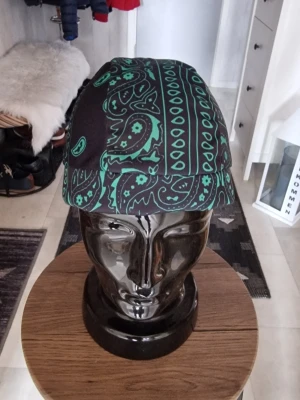 Svart och grön paisleymönstrad bandanas - Snygg bandanas i svart tyg med grönt paisleymönster och klassiska band att knyta baktill. Materialet ser ut att vara mjukt och glansigt, troligen polyester eller satin. Perfekt accessoar för att lyfta din streetwear-look. Bandana pirathatt.  Stretchig snabbtorkande andningsbar och  vindtät. 55-60 cm huvud.