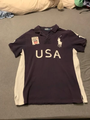 Mörkblå Ralph Lauren USA piké - Snygg mörkblå pikétröja från Ralph Lauren med vita detaljer på sidorna och stora tryckta bokstäver 'USA' framtill. Klassisk polokrage, broderad logga och patch på bröstet. Perfekt för dig som gillar sportig stil.