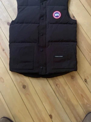 Svart dunväst från Canada Goose - Snygg svart dunväst från Canada Goose med klassisk logotyp på bröstet och två stora framfickor. Västen har en rak passform, tryckknappar framtill och är vadderad för extra värme. Perfekt för lager-på-lager och streetwear-stil.