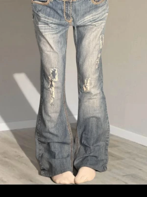 Blå slitna bootcut jeans - Säljer ett par blå bootcut jeans med slitningar och råa detaljer framtill. Jeansen har låg midja, klassiska fem fickor och kontrastsömmar. Perfekta för en avslappnad och trendig look.