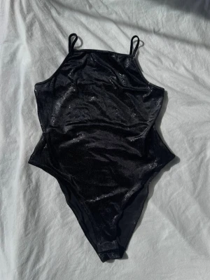 Svart glittrig body från H&M Divided - Snygg svart body med glittrigt tyg och tunna axelband från H&M Divided. Kroppsnära passform och hög skärning vid benen ger en trendig siluett. Perfekt för dig som vill ha något extra till din outfit. Materialet är stretchigt och bekvämt.