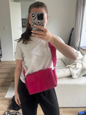 Rosa axelväska med rutmönster - Säljer en snygg axelväska i starkt rosa färg med rutmönstrad design. Väskan har bred axelrem och boxig form, perfekt för att bära det viktigaste. Materialet ser ut att vara konstläder och väskan har en modern vibe.