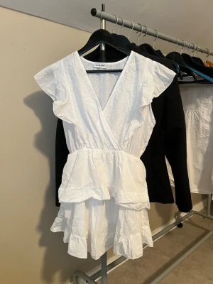 Klänning - Söt vit kortklänning från Meet Me There med omlott-design, v-ringning och volangdetaljer. Klänningen har korta ärmar och ett diskret prickigt mönster i tyget. Perfekt för sommarens alla tillfällen!