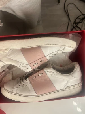 Valentino Garavani sneakers rosa/vit - Snygga sneakers från Valentino Garavani i vitt skinn med bred rosa panel på sidan. Klassisk låg modell med snörning och platt sula. Kommer i originalkartong. Perfekta för dig som vill ha en exklusiv och trendig look. Sulorna i skorna är ej orginala 