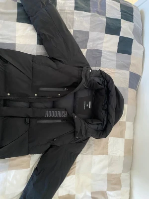 Svart pufferjacka från Hoodrich - KAN DISKUTERA PRIS.       Snygg svart pufferjacka från Hoodrich med huva och dragkedja framtill. Jackan har en cool logga på bröstet och flera praktiska fickor med dragkedja. Perfekt för kalla dagar när du vill hålla stilen och värmen.