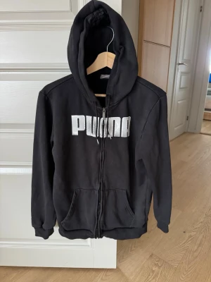 Svart hoodie med dragkedja från Puma - Svart hoodie från Puma med vit logga framtill. Tröjan har dragkedja, huva med snören och två stora fickor på magen. Mjuk bomullsmix och ribbade muddar vid ärmslut och nederkant. Perfekt för en avslappnad och sportig stil. I nyskick!