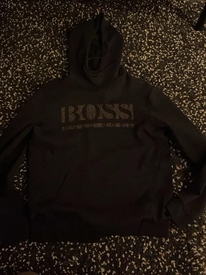 Svart hoodie från BOSS med tryck - Snygg svart hoodie från BOSS med stort guldfärgat BOSS-tryck på bröstet. Klassisk huva och magficka framtill. Perfekt för dig som vill ha en stilren och trendig look. Materialet känns mjukt och bekvämt.