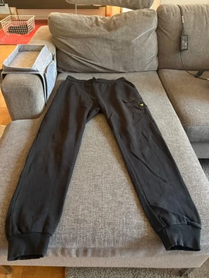 Svarta mjukisbyxor från Lyle & Scott - Säljer ett par svarta mjukisbyxor från Lyle & Scott med resår i midjan och vid bensluten. Byxorna har en ficka på sidan med det klassiska gula örnloggan. Perfekta för chill eller streetwear-stil.