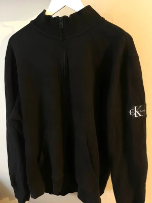 Calvin Klein Jeans quarter zip-tröja - Snygg svart hoodie från Calvin Klein Jeans med mjuk  bomullskvalitet med våfflad struktur, hög krage med dragkedja och klassisk CK-logga på ärmen. Storlek XL
