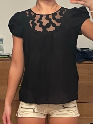 Svart blus med transparent spets - Snygg svart blus med puffärmar och transparent parti upptill dekorerat med spetsdetaljer. Rund halsringning och luftig passform, perfekt för en trendig look. Materialet känns lätt och skönt mot huden.