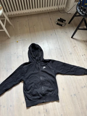 Svart zip hoodie från Nike - Säljer en svart hoodie från Nike med dragkedja framtill och två stora fickor. Klassisk Nike-logga broderad på bröstet. Hoodien har huva med dragsko och är tillverkad i mjukt, fleecefodrat material som är riktigt skönt. Perfekt för chill eller träning.