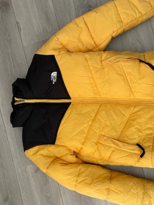 Gul pufferjacka från The North Face - Säljer en riktigt snygg gul pufferjacka från The North Face med svarta detaljer på axlar och krage. Jackan har dragkedja framtill, två sidofickor med dragkedja och logotyp på bröstet och ryggen. Perfekt för kalla vinterdagar och håller dig varm och stilren.