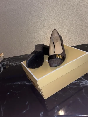 Bruna pumps från Michael Kors - Snygga pumps från Michael Kors i brunt med klassiskt MK-mönster och guldfärgad MK-logga framtill. Skorna har rund tå, låg klack och detaljer i svart läderimitation. Perfekta för dig som vill ha en elegant och trendig look.