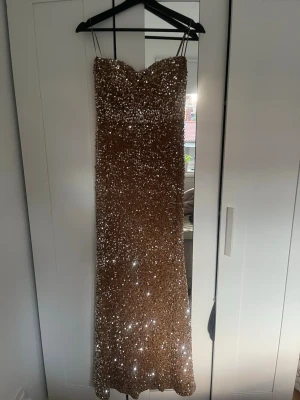Glittrig guld långklänning  - Säljer en superglittrig långklänning i guld som är strapless och  figurnära passform. Klänningen är täckt av paljetter som ger en riktigt lyxig känsla och har en lätt hjärtformad urringning. Perfekt för dig som vill sticka ut på festen! Har även aldrig använt den! 