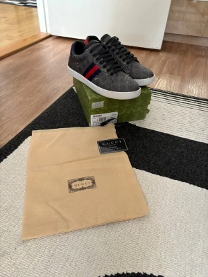 Gucc1 skor med tiger mönster - Snygga Gucc1 sneakers i grått med klassiskt monogrammönster och röda/blå ränder på sidan. Skorna har vita sulor och svarta skosnören. Tillverkade i canvas med läderdetaljer. Perfekta för dig som vill ha en lyxig och trendig look.