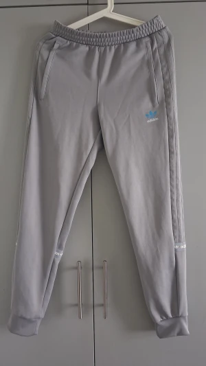 Gråa mjukisbyxor från Adidas - Snygga grå mjukisbyxor från Adidas med elastisk midja och muddar vid bensluten. Byxorna har två sidofickor och klassiska Adidas-loggor i vitt och blått på benet och nedtill. Tillverkade i mjukt material som passar perfekt för chill dagar.