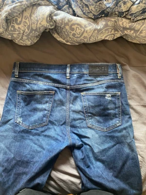 Blå slitna jeans från Diesel - Säljer ett par blå jeans från Diesel med snygga slitningar och klassisk femficksdesign. Jeansen har en mörk tvätt med ljusare partier och detaljerade slitningar på både fram- och baksida. Perfekta för en avslappnad och trendig look.
