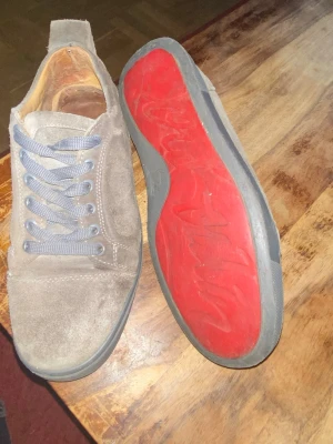Christian Louboutinu - Snygga sneakers i beige mocka med grå skosnören och en cool röd yttersula. Skorna har en klassisk låg profil och rund tå, vilket ger en clean och stilren look. Perfekta för dig som gillar en enkel men ändå unik stil. Kan gå med på billigare priser vid snabba affärer 